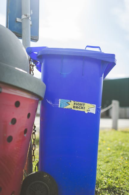 A tall, rectangular blue outdoor waste bin with a closed lid, positioned on a grassy area next to a pink wheelie bin with black ventilation holes and a partially visible gray lid. The blue bin has a y
