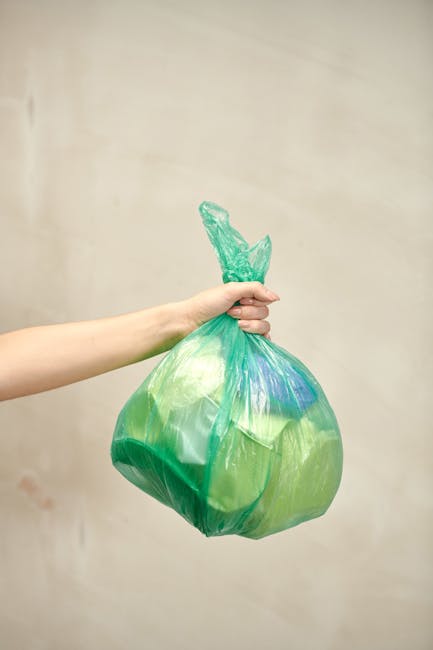A person's hand and forearm extend from the left side of the image, holding a green plastic rubbish bag by its tied top. The bag appears to contain mixed waste materials and has a slightly crinkled te