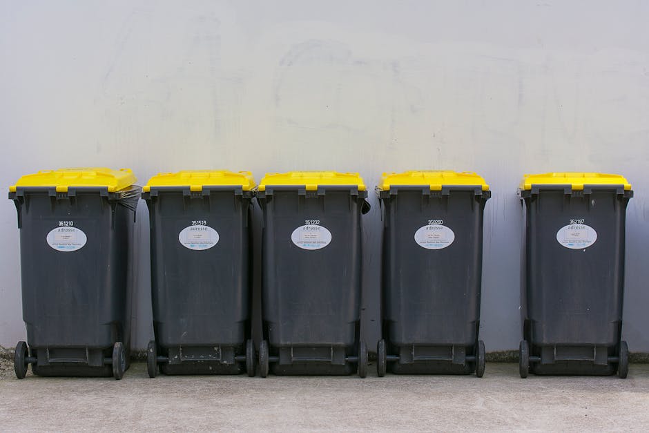 A large, weathered metal rubbish disposal container positioned outdoors on a paved surface, with a sloped top and rust streaks visible along its edges. The container features a prominent sign with the